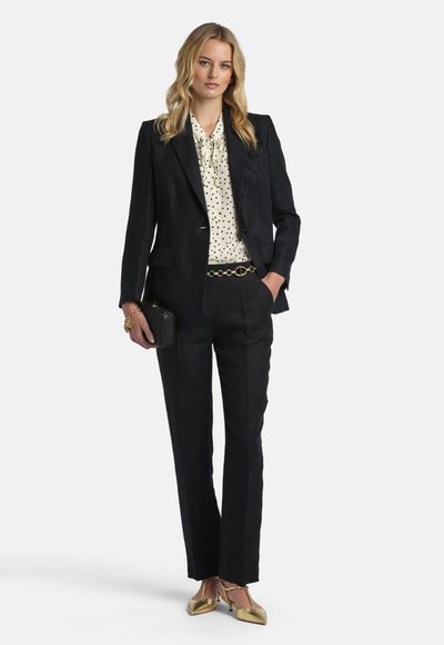 Woman wearing black tailored suit with polka dot blouse, gold belt, gold heels, carrying black clutch, standing with one hand in pocket.