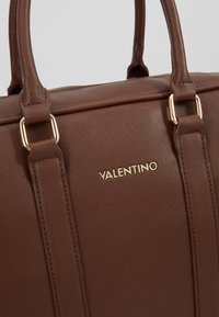 Brun strukturerad läder tote-väska med detaljer i guldton, med dubbla handtag och en logotyp på framsidan.