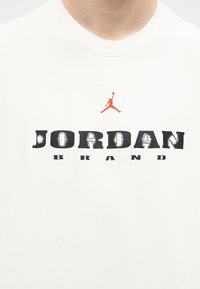 Λευκό πουκάμισο με μαύρο κείμενο "JORDAN BRAND" και μικρή πορτοκαλί σιλουέτα ενός μπασκετμπολίστα που πηδάει με τη μπάλα πάνω από το κείμενο.