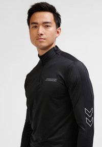 Schwarzes langärmliges Sportoberteil mit einem halb offenen Kragen, das ein dezentes Logo auf der Brust und Chevron-Akzente an den Ärmeln aufweist.