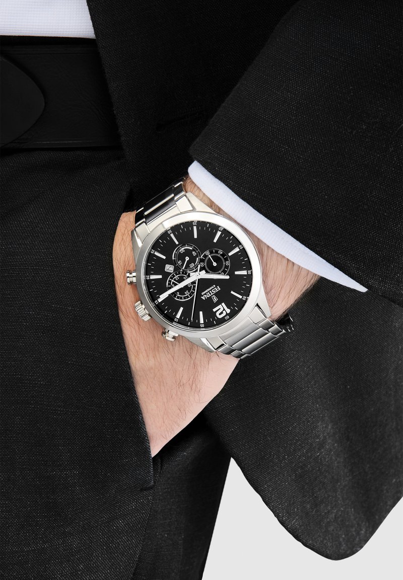 Silberner Edelstahl-Uhren mit schwarzem Zifferblatt, Chronographen-Unterzifferblättern und Datumsanzeige. Getragen an einem Handgelenk mit einem schwarzen Sakko.