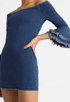 Off-shoulder tætsiddende blå denimkjole med lagdelte frynseklokkeærmer og rå kanter, båret af en person med hånden på hoften.