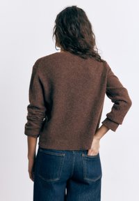 Žena s kovrčavom tamnom kosom nosi smeđi pleteni džemper i plave traperice, stoji s jednom rukom u stražnjem džepu, okrenuta leđima.