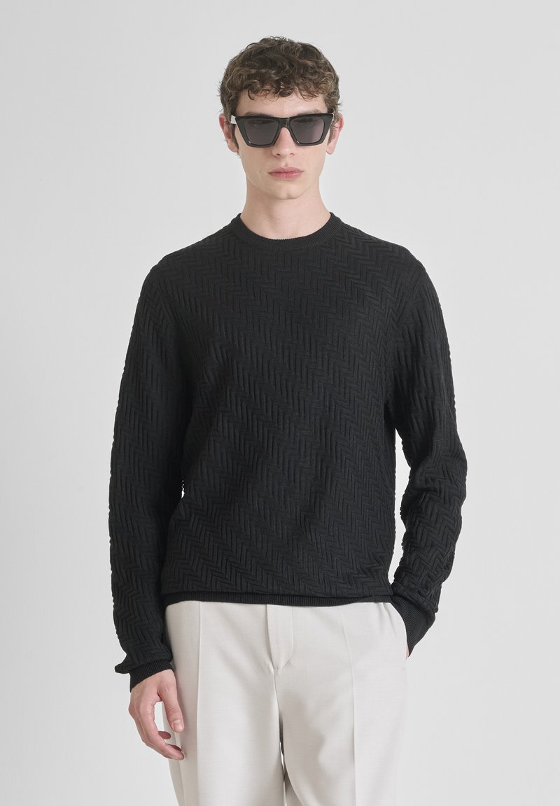 Zwarte gestructureerde trui met een zigzagpatroon, ronde halslijn en lange mouwen, gecombineerd met lichtgekleurde broeken en donkere zonnebril.