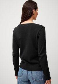 Maglione nero lavorato a maglia con scollo sulla schiena, maniche lunghe e polsini e orlo a costine. Indossato con jeans blu a vita alta.
