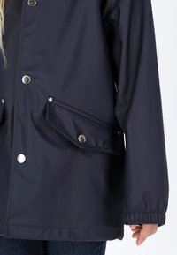 Giacca impermeabile blu navy realizzata in materiale liscio; presenta bottoni a pressione, due tasche frontali inclinate e polsini elastici per una vestibilità aderente.