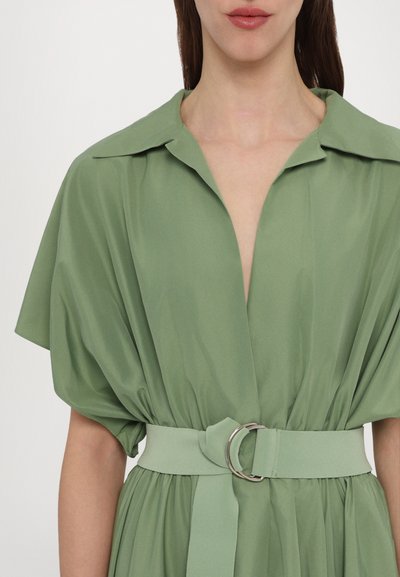 Vestido verde com gola e mangas curtas drapeadas. Apresenta uma cintura ajustada com um cinto combinando e uma fivela de anel prateada.