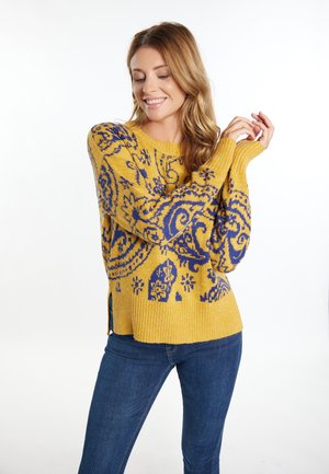 Yellow knitted sweater featuring a blue paisley pattern, ribbed texture, and side slits. Paired with blue jeans.