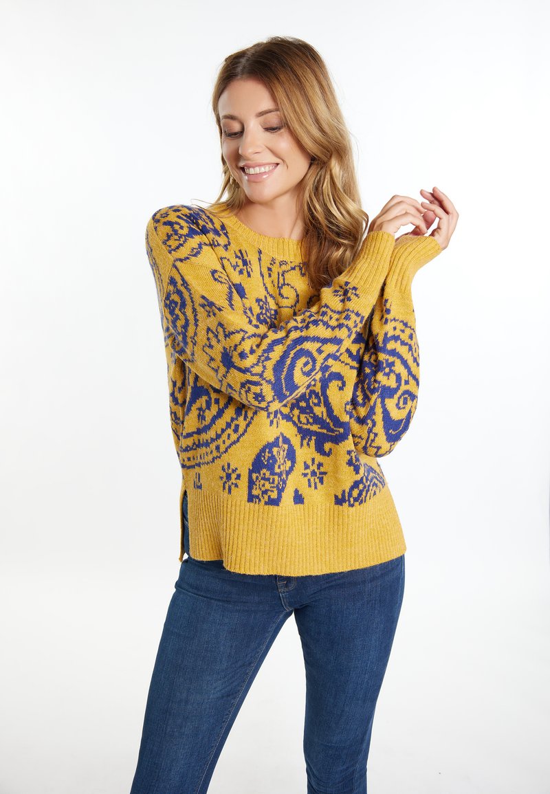 Geel gebreide trui met een blauw paisleypatroon, ribbeltextuur en zijsplitten. Gecombineerd met blauwe jeans.