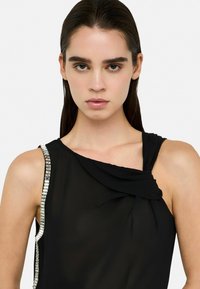 Black sleeveless top with draped front, featuring a sleek design and a silver sequin accent on the left shoulder. Soft, smooth fabric texture.