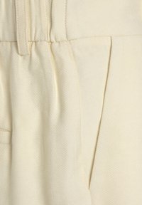 Tessuto texturizzato crema con pieghe, cinta in vita e tasca laterale, che enfatizza un design liscio e strutturato.