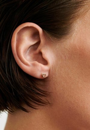 Close-up af en persons øre med kort brunt hår iført en lille guldfarvet ørestik i form af et hjerte.
