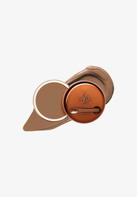 Produit de maquillage compact avec un couvercle en bronze circulaire, une crème brun clair à l'intérieur, et un échantillon lisse de teinte beige à proximité.