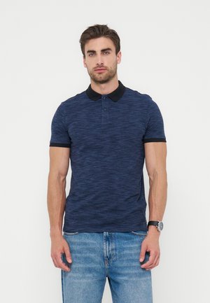 Muž oblečený v tmavomodrom polotrčku s čiernym golierom a lemovaním rukávov, svetlomodrými džínsy a náramkovými hodinkami, stojaci pred bielym pozadím.