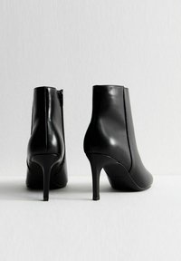 Botas de cuero negras hasta el tobillo con un diseño elegante, punta afilada, tacón alto de aguja y detalle de cremallera lateral. Textura suave con acentos mínimos.