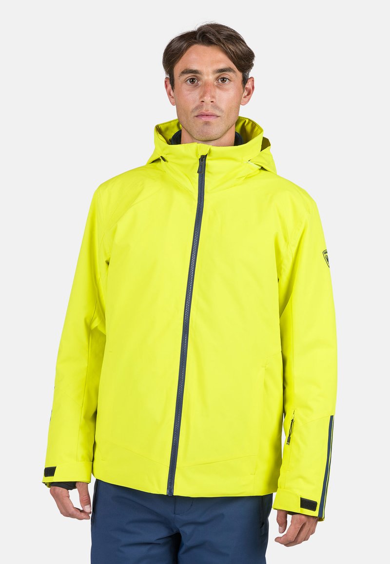 Chaqueta impermeable amarilla brillante con capucha, que cuenta con un cierre frontal y bolsillos laterales. Textura suave con acentos en azul marino en contraste.