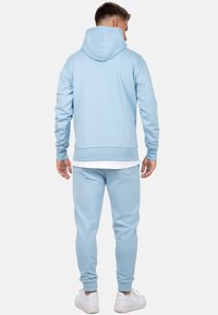 Lichtblauwe hoodie en bijpassende joggers, gemaakt van zacht materiaal. Heeft een capuchon, ribgebreide manchetten en een elastische tailleband. Draagt met witte sneakers.
