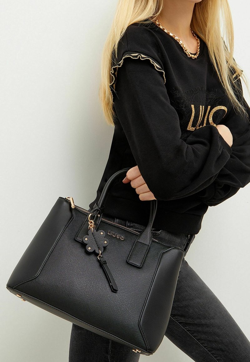 Femme aux longs cheveux blonds portant un pull noir et un jean, tenant un sac à main en cuir noir texturé avec un charm et des détails dorés.