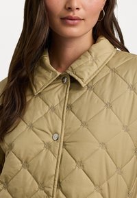 Pukinė bežinė striukė su apykakle, mygtukų uždarymu ir siuvinėtų logotipų akcentais. Lygus sintetinis audinys su deimanto raštu.