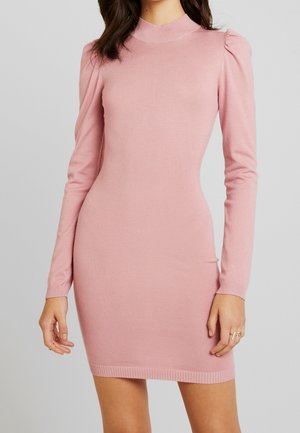 Femme portant une robe en maille rose ajustée à manches longues avec un col montant et des épaules légèrement bouffantes, sur un fond gris uni.