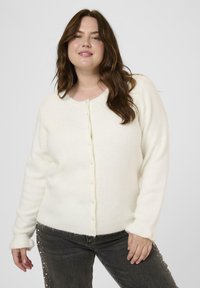 Smiling woman with long brown hair wears a white buttoned cardigan and black jeans adorned with pearl details on the sides.