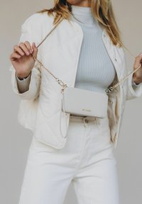 Sac en cuir gris clair à bandoulière, avec une fermeture éclair, surface texturée, quincaillerie dorée et une chaîne amovible, porté sur une veste blanche matelassée.