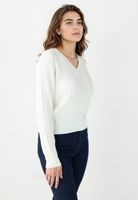 V-neck white knitted sweater with a textured cable pattern, ribbed cuffs and hem, paired with dark blue slim-fit jeans.