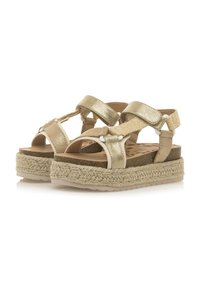 Sandalias en tono dorado con tiras cruzadas, que presentan una plataforma de yute trenzado y una plantilla en tono beige claro. Materiales lisos y texturizados contrastan en el diseño.
