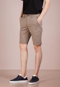 Shorts kaki avec un ourlet roulé, en toile de coton, dotés de poches latérales et de passants de ceinture. Portés avec des baskets noires.