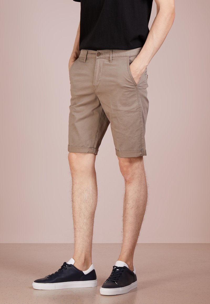 Shorts kaki avec un ourlet roulé, en toile de coton, dotés de poches latérales et de passants de ceinture. Portés avec des baskets noires.
