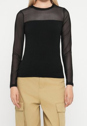 Frau trägt ein schwarzes Langarm-Top mit transparentem Mesh am oberen Teil und an den Ärmeln, kombiniert mit beige-farbenen Cargohosen mit Klappentaschen.