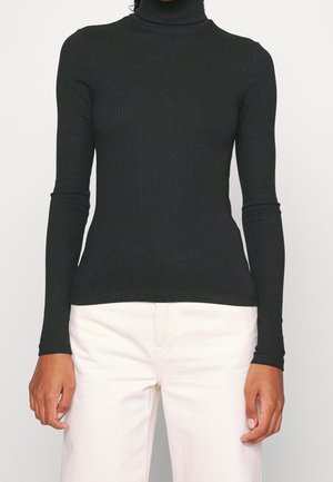 Czarny prążkowany golf z długim rękawem noszony z wysokim stanem białych spodni, ręce swobodnie opuszczone wzdłuż ciała na jednolitym tle.