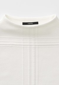Tessuto bianco con texture, colletto alto e sottili motivi a strisce orizzontali e verticali in rilievo. Etichetta nera con testo bianco "someday."