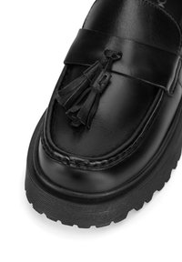 Černé kožené mokasíny s třásňovou ozdobou, které mají silnou, strukturovanou gumovou podrážku a podrobné prošívání kolem špičky.