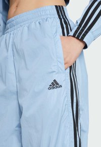 Ljust blå träningbyxor med elastisk midja, med svarta sidostripes och en broderad svart Adidas-logotyp på vänster höftficka.