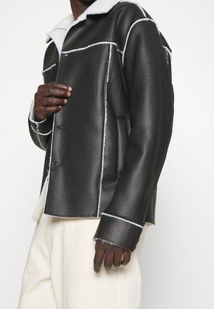 Homme portant une veste en cuir noir avec doublure en shearling blanche et un pantalon blanc, ajustant l'avant de la veste devant un fond blanc.