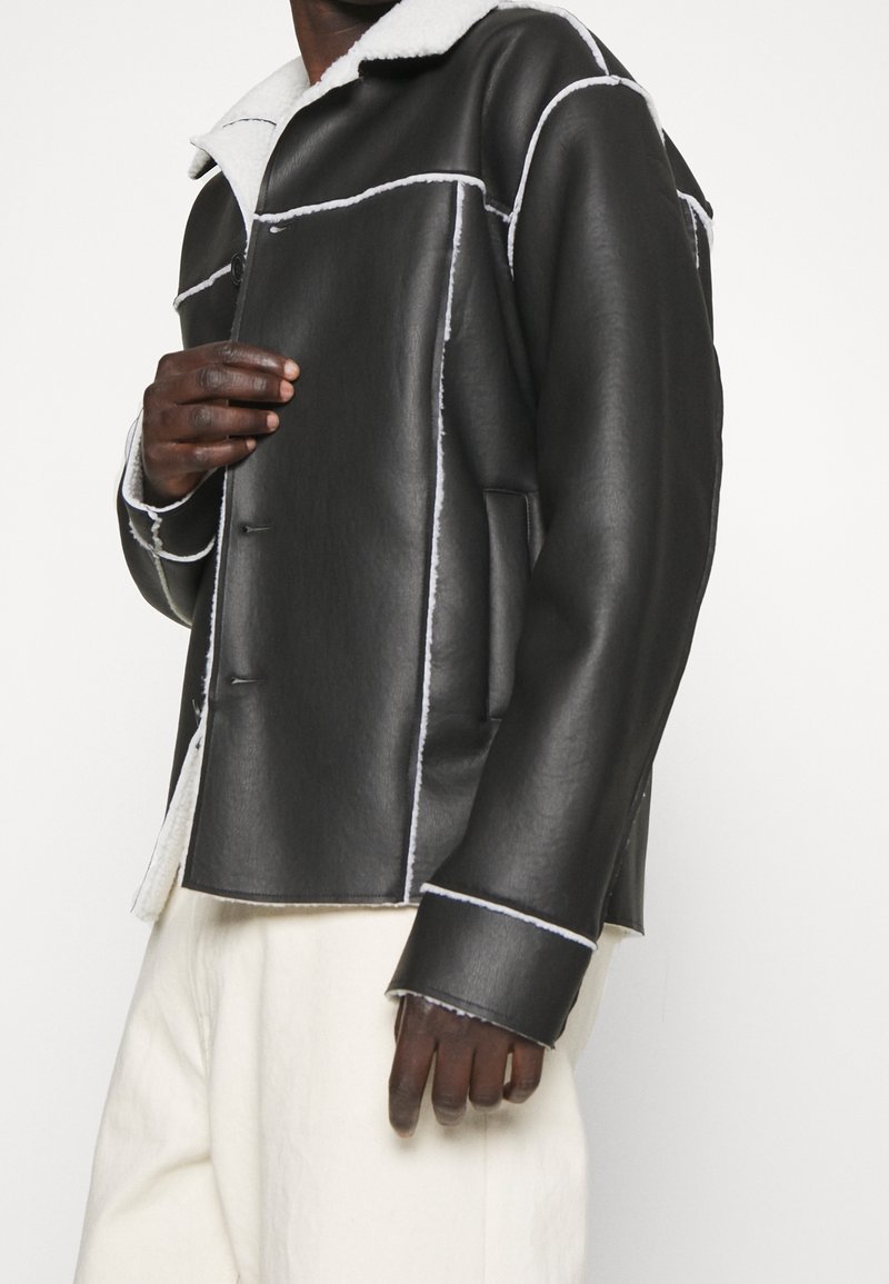 Uomo con giacca di pelle nera con foderatura in shearling bianco e pantaloni bianchi, che sistema la parte davanti della giacca contro uno sfondo bianco.