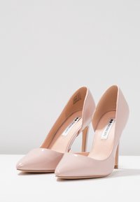 Beige lakované lodičky s špičatou špičkou a štíhlým jehlovým podpatkem. Hladký povrch a minimalistický design bez ozdob.