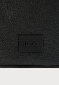 Must tekstureeritud nahast kott, millel on ristkülikukujuline märk, millel on musta värvi reljeefne brändinimi "HUGO". Lihtne disain peene detailiga.