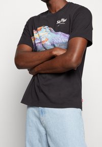 Svart bomullströja med en färgglad bergsgrafik och logotyp framtill, med en klassisk rund hals och korta ärmar.