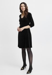 Schwarzes Samtkleid mit V-Ausschnitt und dreiviertel Ärmeln. Abgerundet mit einem Taillenbund und einem fließenden Rock. Tragen mit schwarzen High Heels.