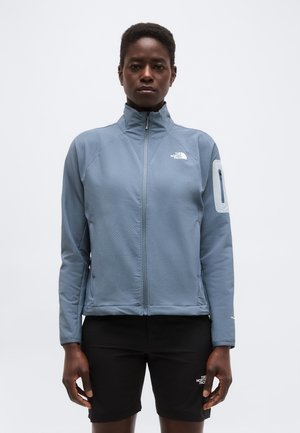 Čierny človek oblečený v svetlomodrej bunde na zips a čiernych šortkách, stojaci pred jednoduchým bielym pozadím.