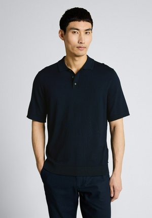 Mężczyzna stojący z rękami w kieszeniach, ubrany w czarną koszulkę polo z krótkim rękawem i ciemne spodnie na jednolitym jasnym tle.