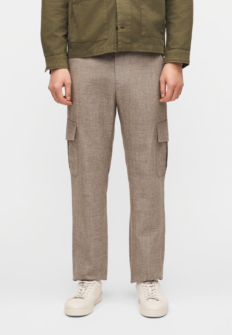 Άνδρας με μπεζ παντελόνι cargo με πλαϊνές τσέπες, λευκά αθλητικά παπούτσια και χακί πράσινο μπουφάν, στέκεται μπροστά σε απλό φόντο.