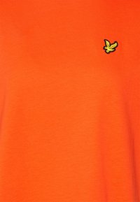 Lys orange stof med et lille broderet gult fuglelogo øverst til højre.