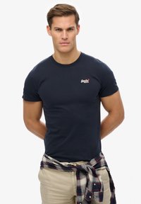 Camiseta de manga corta azul marino hecha de algodón, con un pequeño logo en el pecho y un diseño sencillo. Llevada con una camisa a cuadros atada a la cintura.