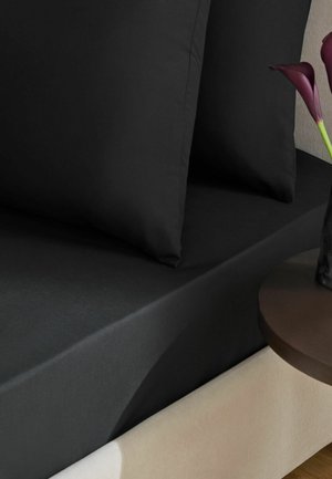 Schwarze Bettwäsche und Kissen auf einem beigen Bettrahmen mit einem kleinen runden Tisch, der eine dunkle Vase mit dunkelvioletten Calla-Lilien hält.