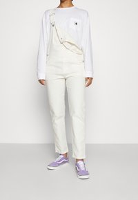Salopette in denim bianco con spalline regolabili, tasca frontale e vestibilità ampia, abbinata a una maglietta bianca a maniche lunghe e a scarpe da ginnastica lavanda.