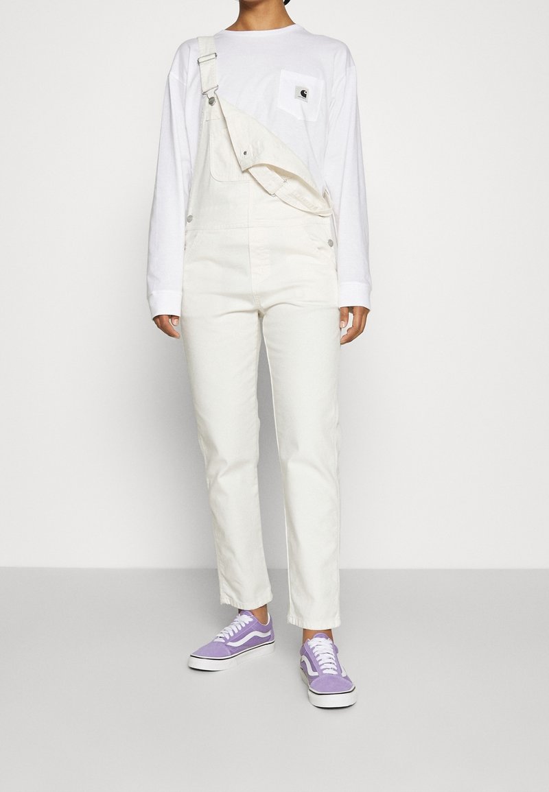 Salopette in denim bianco con spalline regolabili, tasca frontale e vestibilità ampia, abbinata a una maglietta bianca a maniche lunghe e a scarpe da ginnastica lavanda.