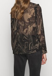 Vrouw met lang bruin haar, gekleed in een doorschijnende blouse met lange mouwen met een bruin en zwart marmerpatroon, gecombineerd met donkere spijkerbroek.